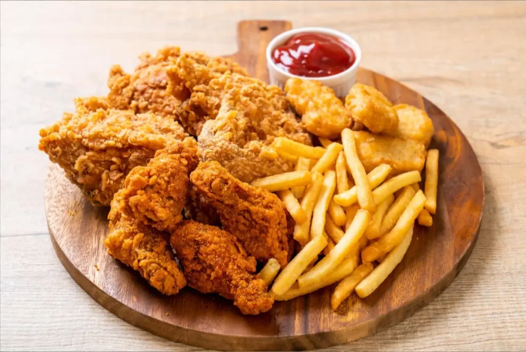 A wooden serving board with crispy fried chicken, spicy wings, golden French fries, nuggets, and a small cup of ketchup.