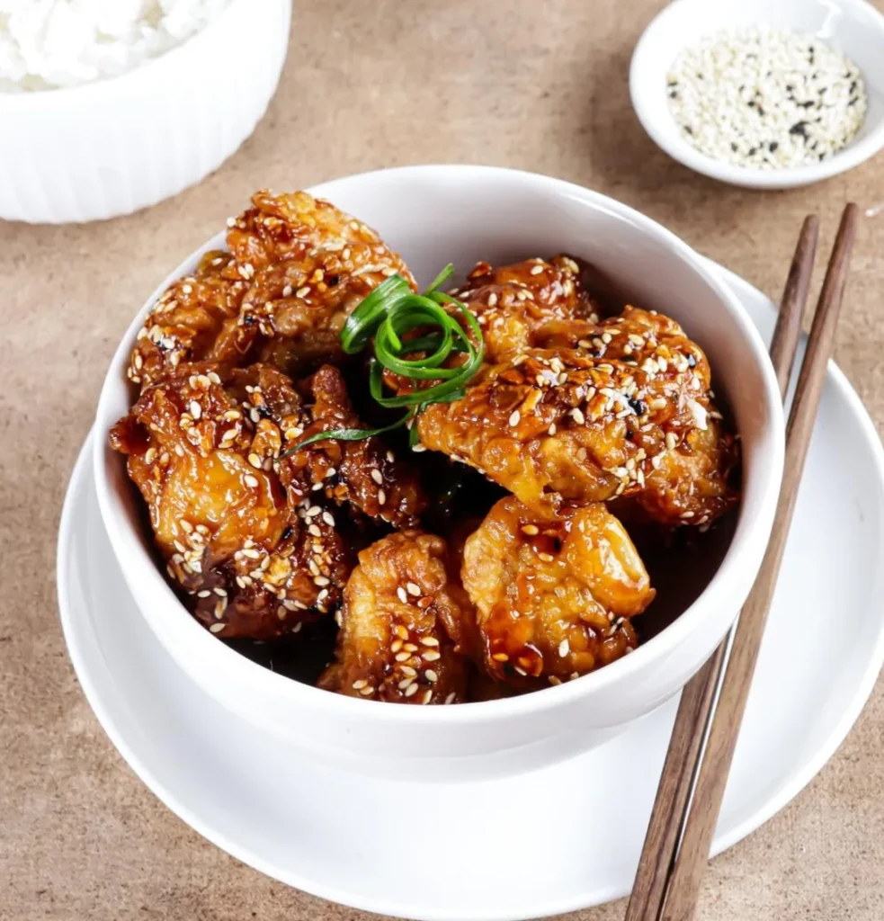 Crispy air fryer teriyaki chicken in a white bowl, garnished with sesame seeds and green onions, served with rice and chopsticks.