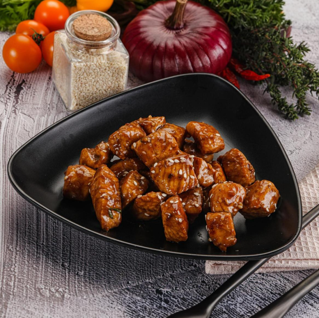 Bite-sized teriyaki chicken pieces on a black plate, garnished with sesame seeds, with fresh ingredients in the background.