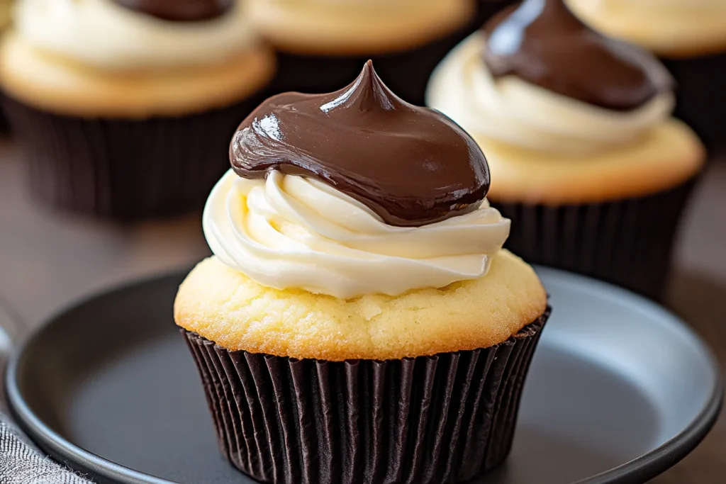 A close-up of a Boston Cream Pie cupcake with vanilla filling, topped with a swirl of whipped cream and glossy chocolate ganache.