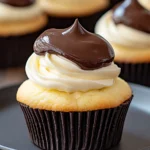 A close-up of a Boston Cream Pie cupcake with vanilla filling, topped with a swirl of whipped cream and glossy chocolate ganache.